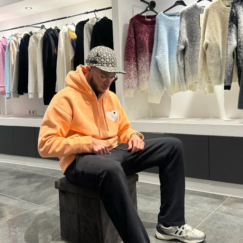 Modèle assis avec le pull ADJ orange clair pour une attitude décontractée mettant en avant le cœur blanc brodé.