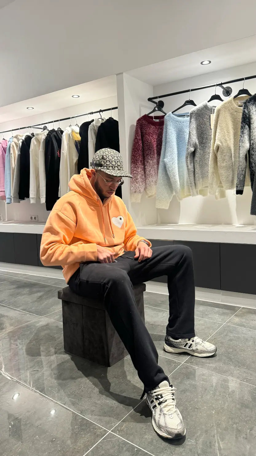 Modèle assis avec le pull ADJ orange clair pour une attitude décontractée mettant en avant le cœur blanc brodé.