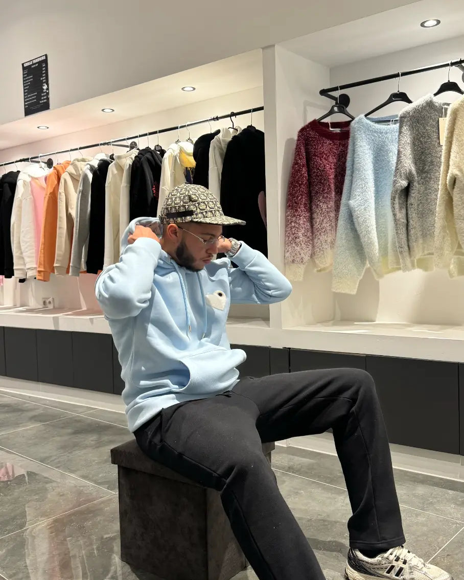 Modèle assis avec le pull ADJ bleu clair et son cœur blanc brodé pour un look streetwear décontracté.