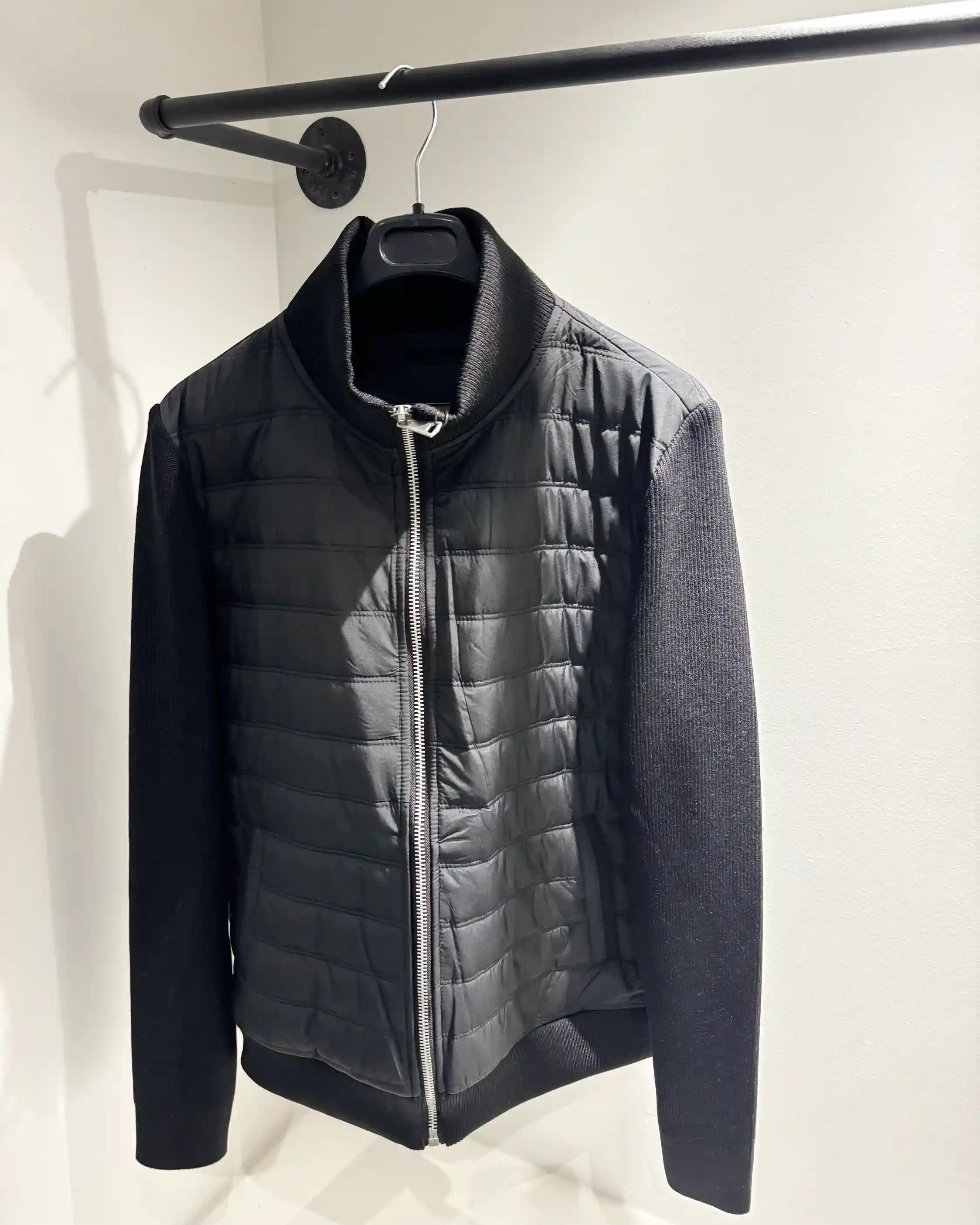 Présentation de la veste hybride noire sur cintre, illustrant le mélange des matières et le tombé impeccable du modèle.