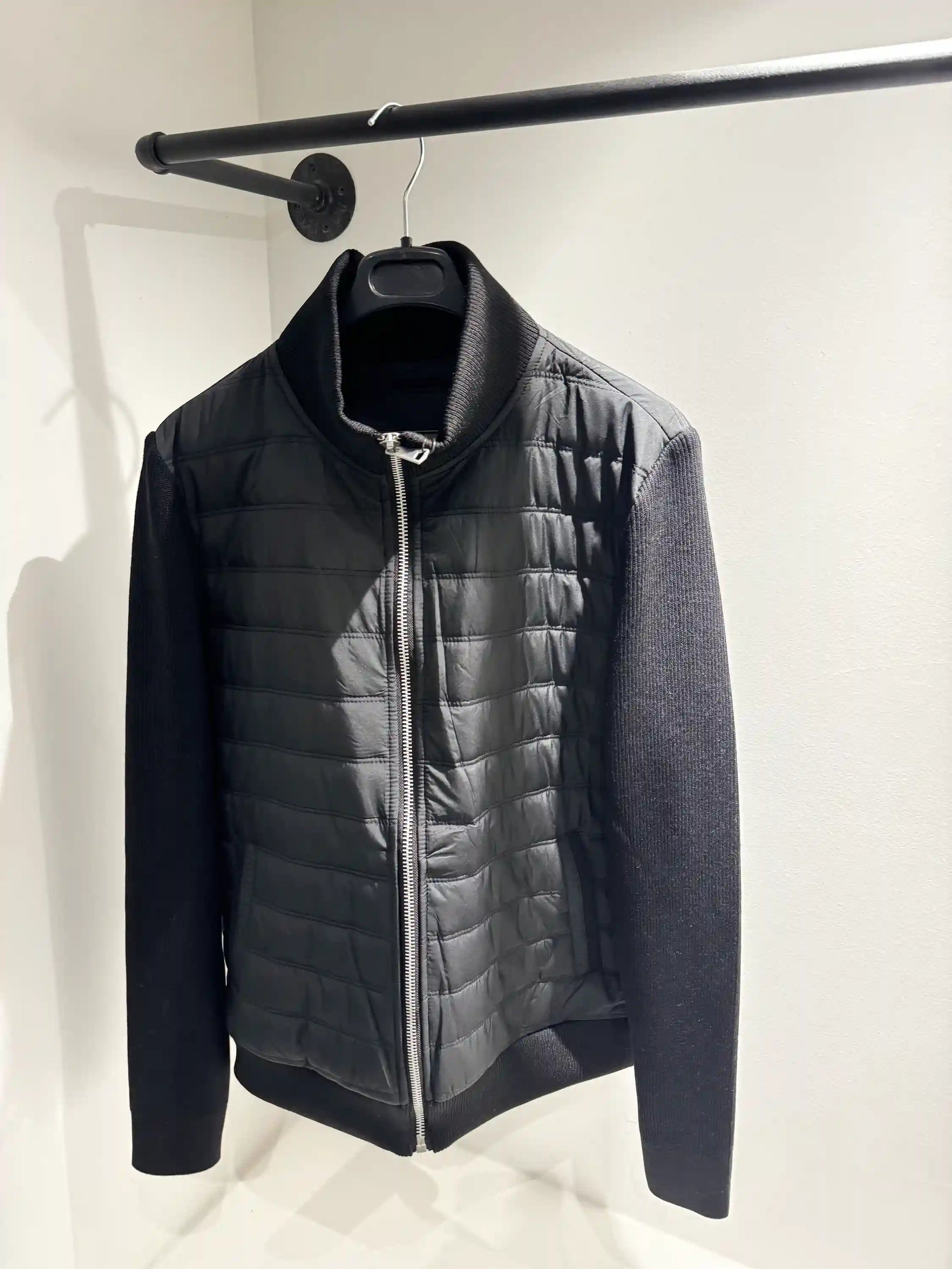 Présentation de la veste hybride noire sur cintre, illustrant le mélange des matières et le tombé impeccable du modèle.